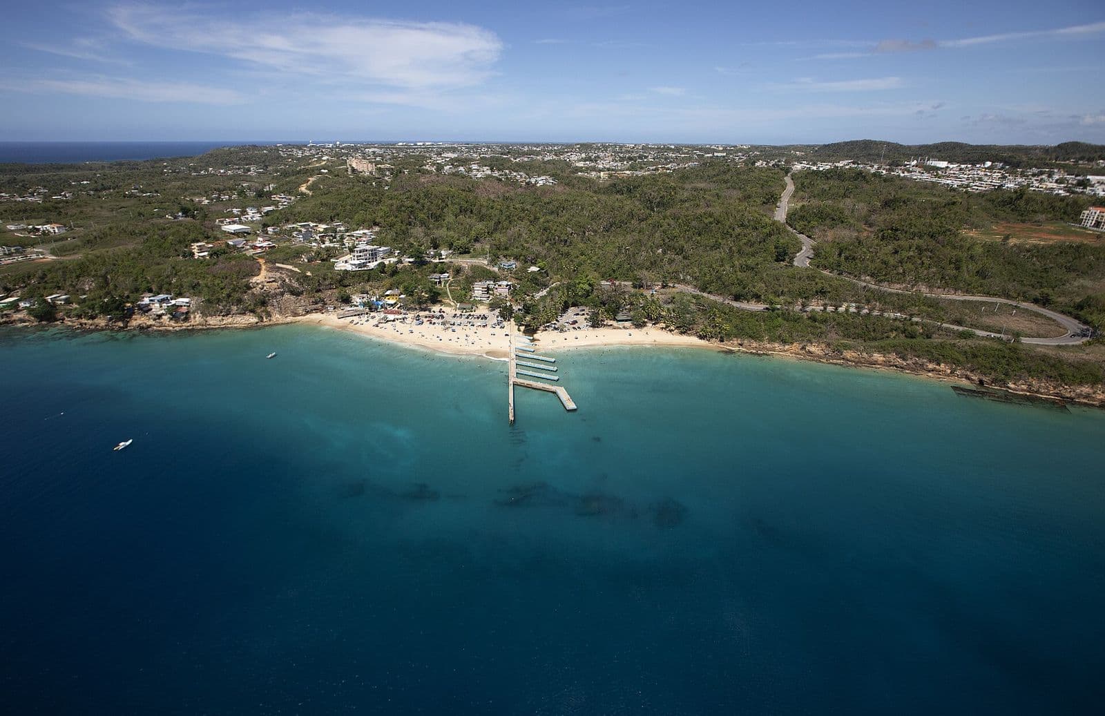 Aerial view of Crash Boat beach in Aguadilla: its long pier reaching into turquoise water, the sand beach and wooded hills, with the town spread across the plateau behind