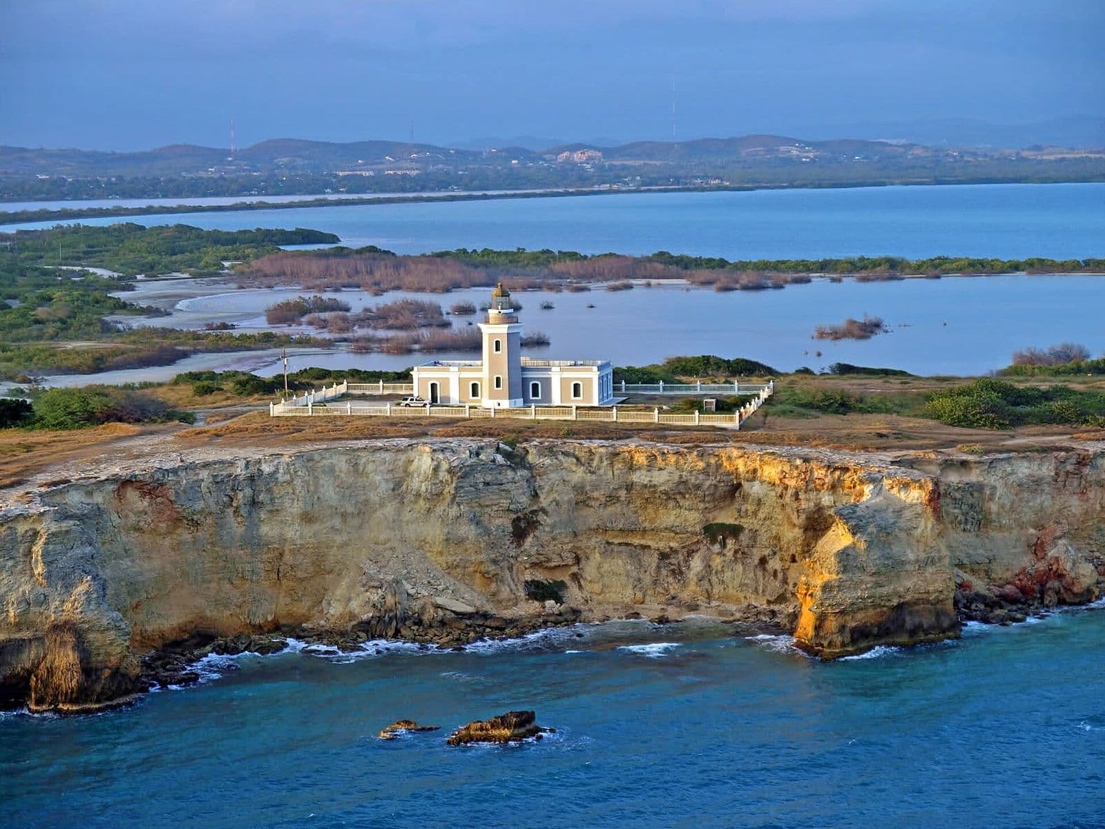The Los Morrillos lighthouse and its white fence on top of tall, sunlit cliffs at Cabo Rojo’s southwest tip, with lagoons, mangroves and hills behind
