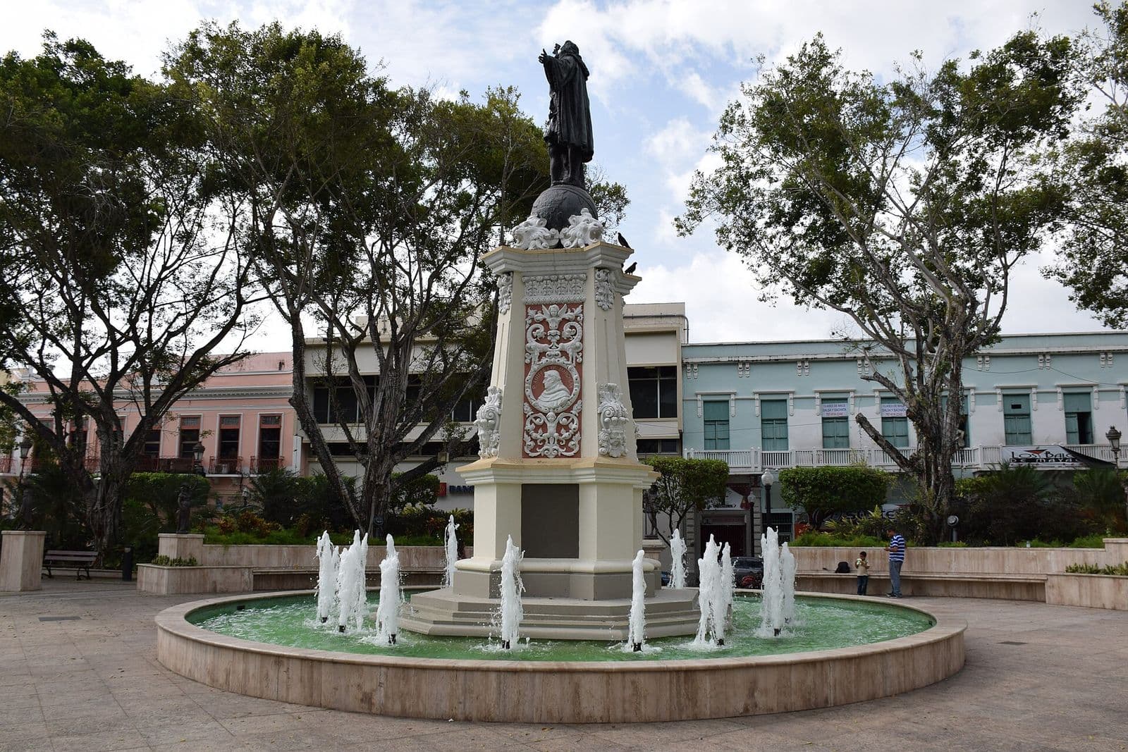 The Christopher Columbus statue and its fountain in the tree-shaded Plaza de Colón, Mayagüez, with pastel two-story buildings around the square