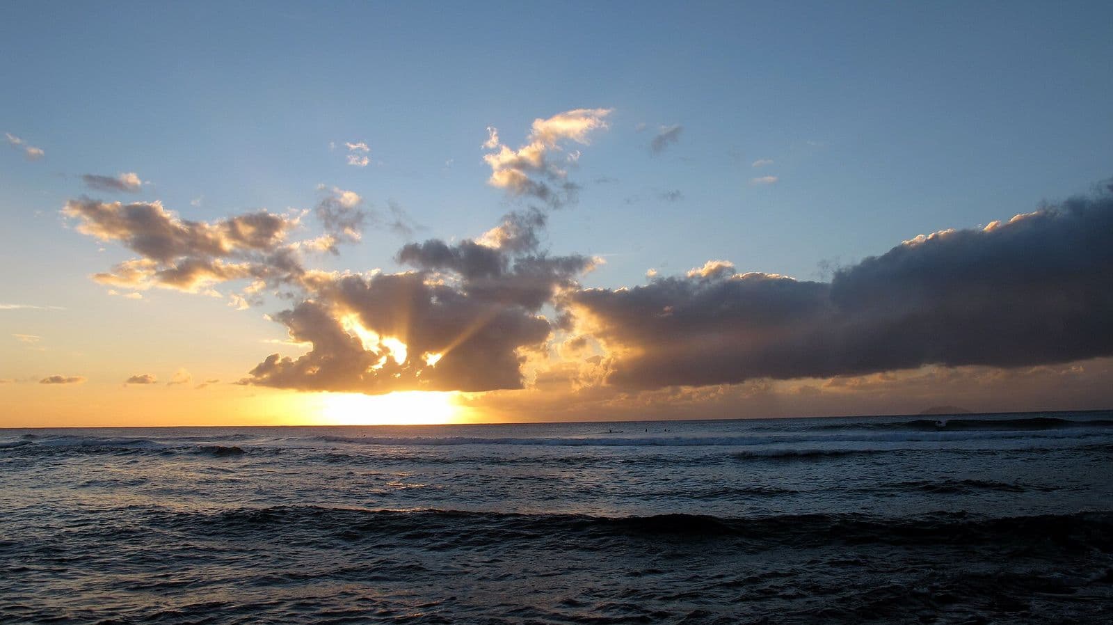 The sun sets behind broken clouds over the sea at María’s beach in Rincón, with surfers waiting in the lineup and Desecheo Island on the horizon