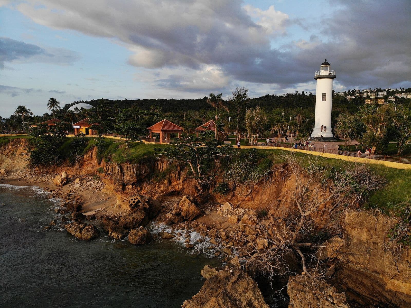 The white Punta Higüero lighthouse in Rincón at golden hour, above eroded cliffs and rocks, with the park’s tile-roofed kiosks and hillside homes behind