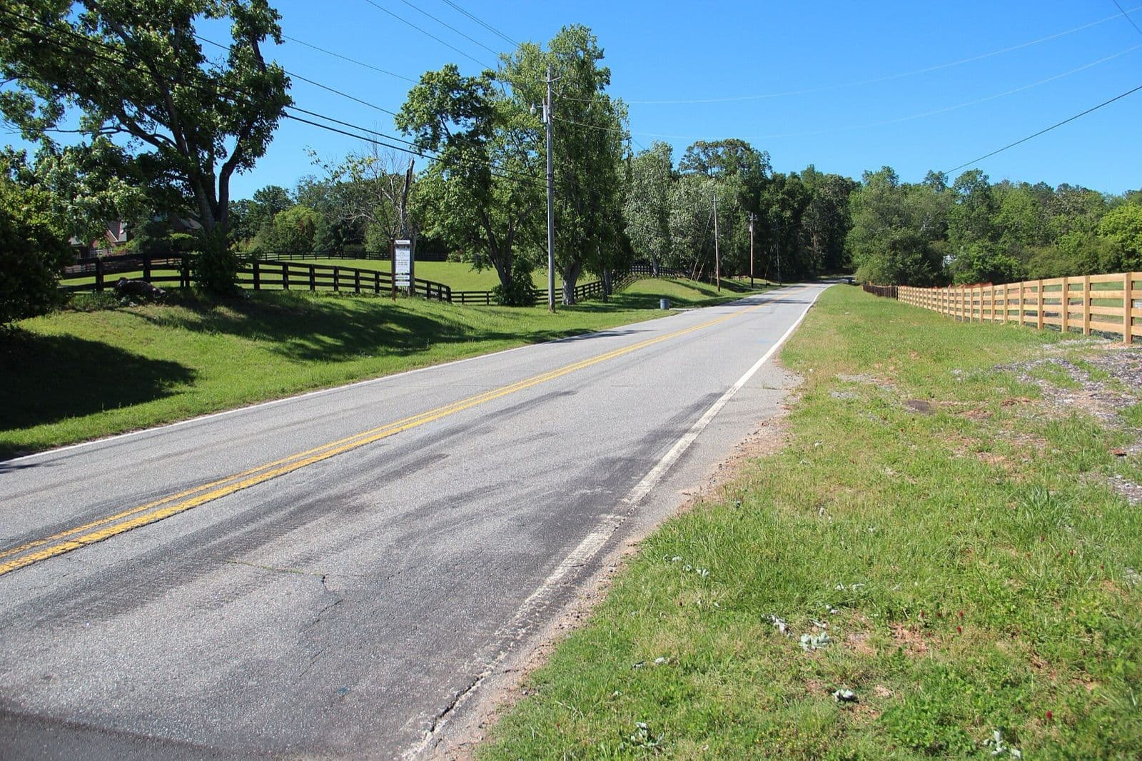 A two-lane country road in Milton between black and natural wood horse-pasture fences, green fields and big shade trees