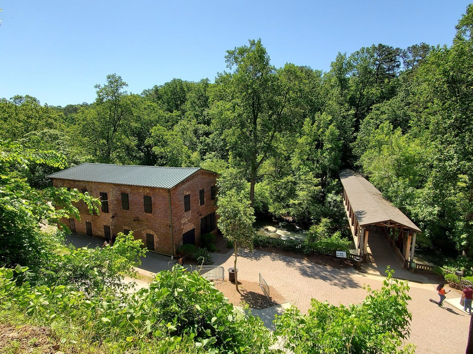 The brick Roswell Mill building and the covered footbridge over Vickery Creek, seen from above among green summer trees