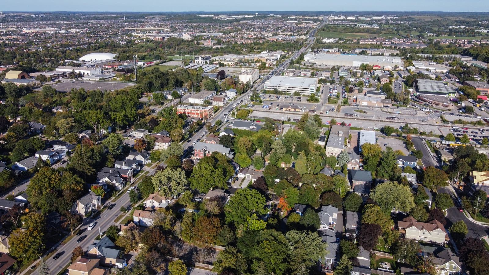 Aerial view over Aurora along Wellington Street: older houses under mature trees in the foreground, a rail line, parking lots and low commercial buildings in the middle, and newer subdivisions to the horizon
