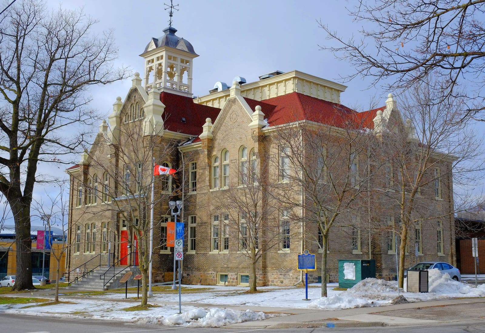 The restored yellow-brick Church Street School in Aurora, with a red roof, an ornate white cupola and a Canadian flag, on a sunny winter day with snow on the lawn