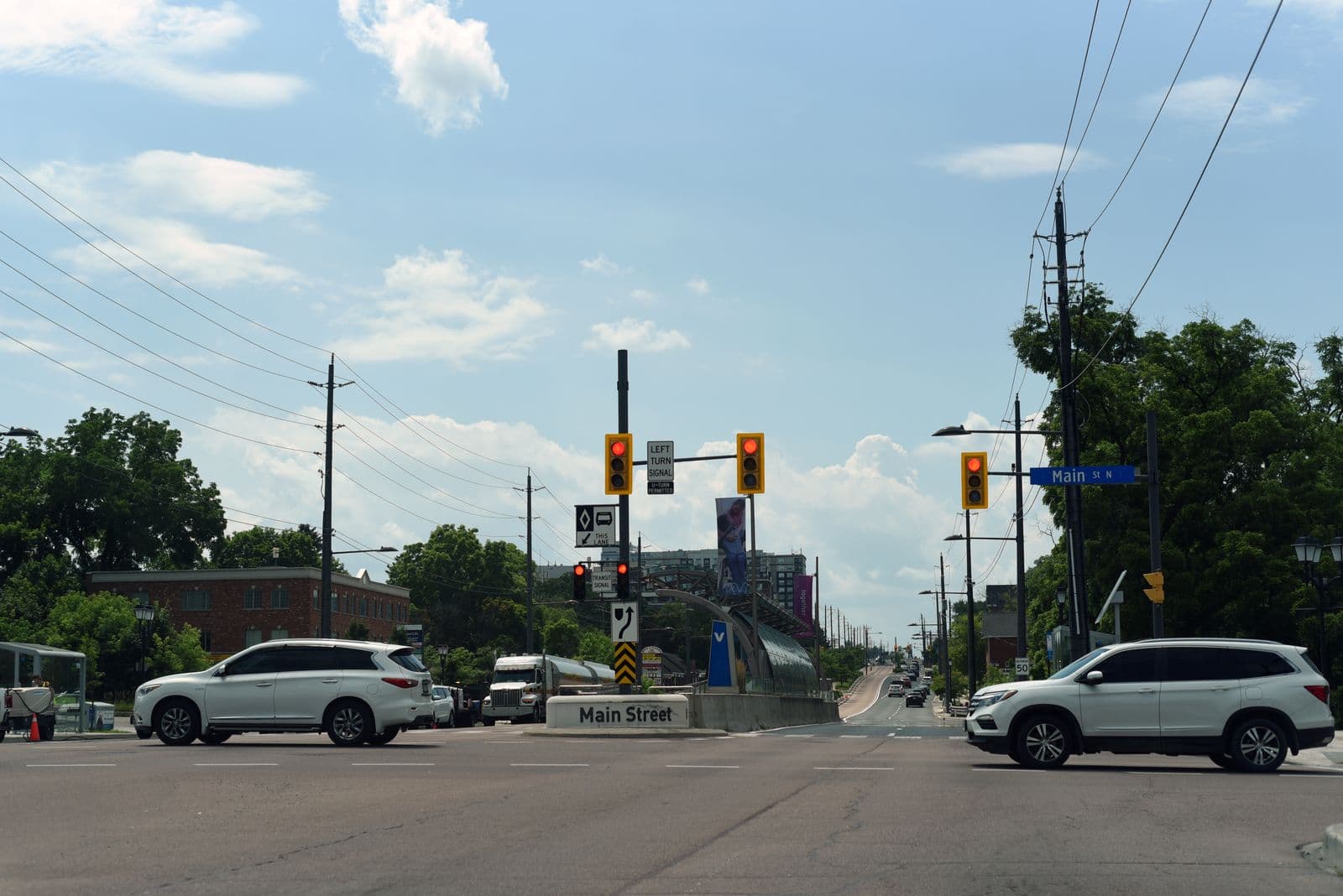 The Main Street vivastation in the middle of Davis Drive in Newmarket, a curved glass shelter between the bus lanes of the rapidway, at a signalled intersection with cars and trees