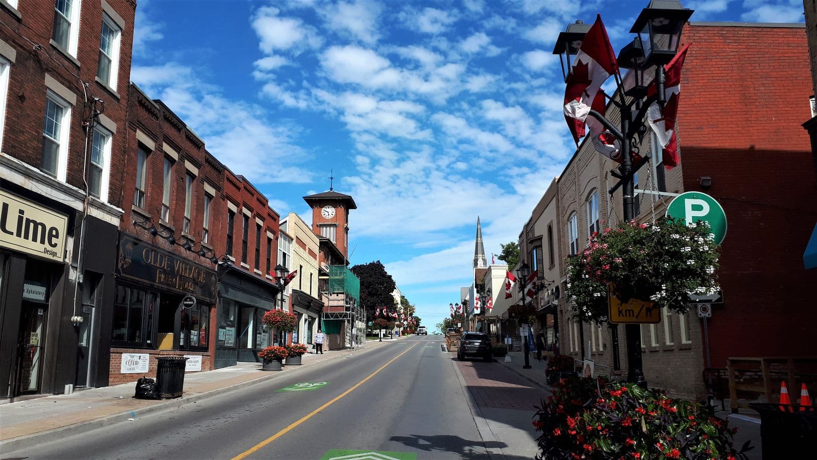 Newmarket’s Main Street lined with two- and three-storey brick storefronts, hanging flower baskets and Canadian flags, with a brick clock tower and a church spire ahead under a blue sky
