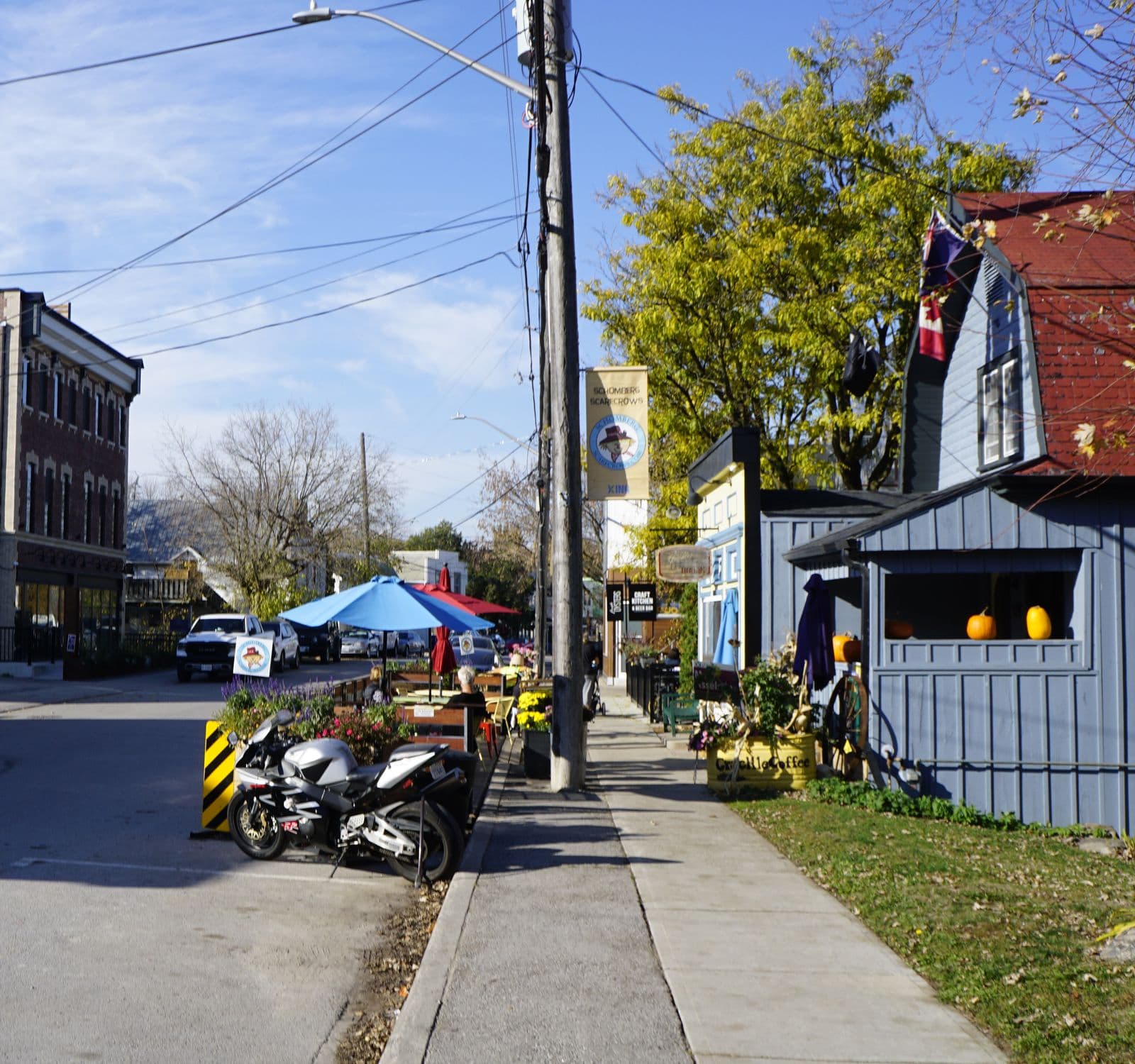 Schomberg’s Main Street on a sunny autumn day, with café umbrellas, flower planters and a blue clapboard shop decorated with pumpkins beside the sidewalk