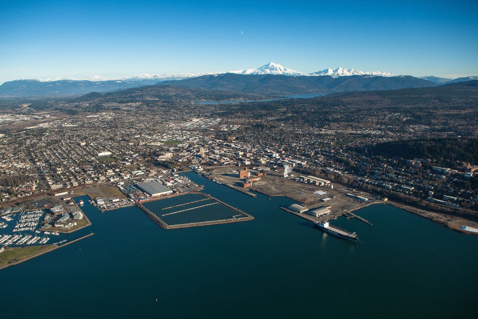 Aerial view across Bellingham Bay to the waterfront and downtown Bellingham, with Lake Whatcom and snow-capped Mount Baker on the horizon under a clear blue sky