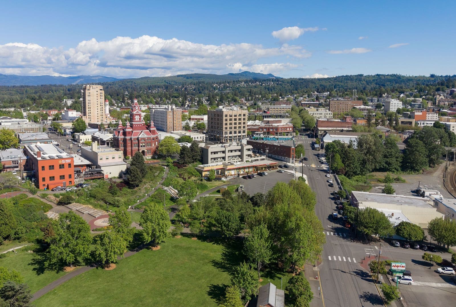 Downtown Bellingham from above on a sunny spring day: a red-brick Victorian building with a white clock tower, office buildings and a green park in the foreground, with forested hills behind