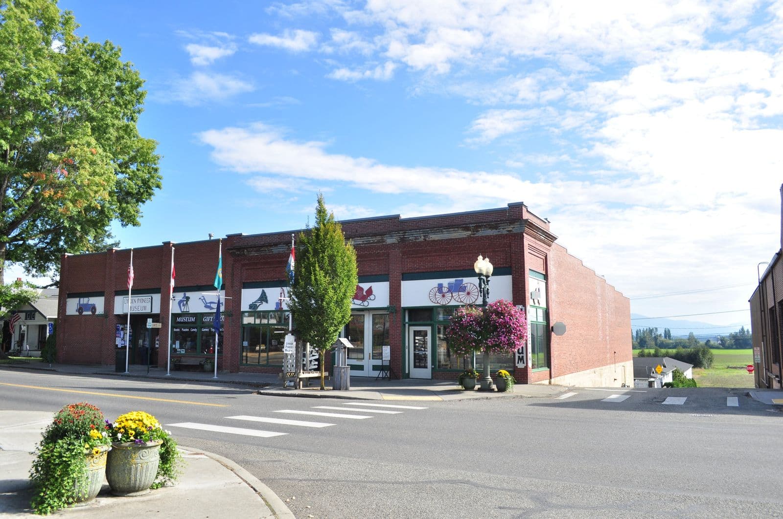 The red-brick Lynden Pioneer Museum on Front Street with flags, hanging flower baskets and planters, and green farm fields beyond the end of the street