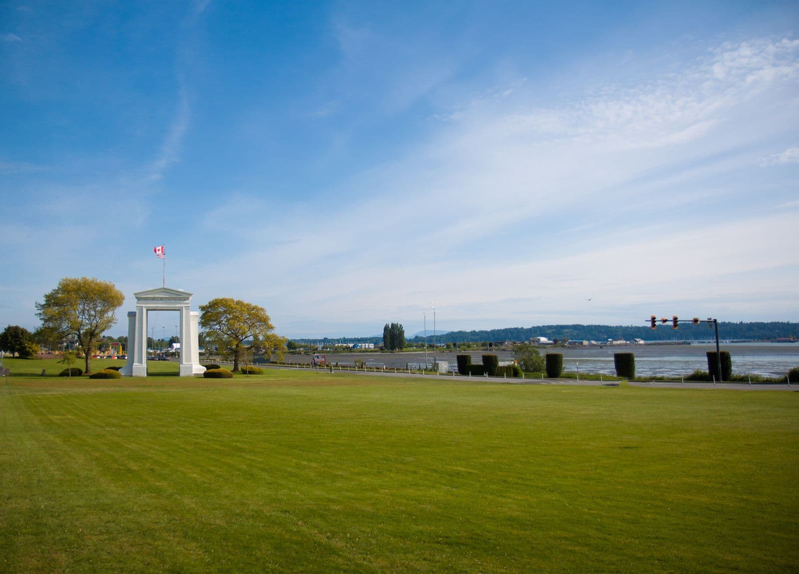 The white Peace Arch on a wide green lawn under a blue sky, with Semiahmoo Bay and the Blaine shoreline to the right
