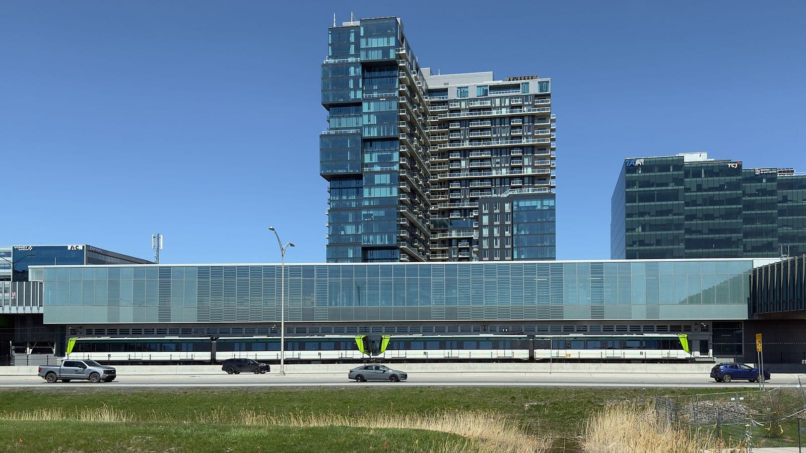 Un train blanc et vert du REM en gare dans la station vitrée Du Quartier, le long de l’autoroute 10 à Brossard, avec les tours de Solar Uniquartier derrière