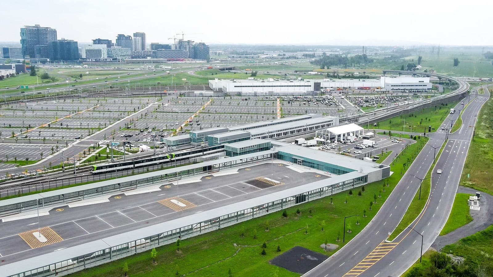 Vue aérienne du terminus Brossard du REM, avec un train à quai, le terminus d’autobus couvert, les stationnements incitatifs et les tours de Solar Uniquartier au loin
