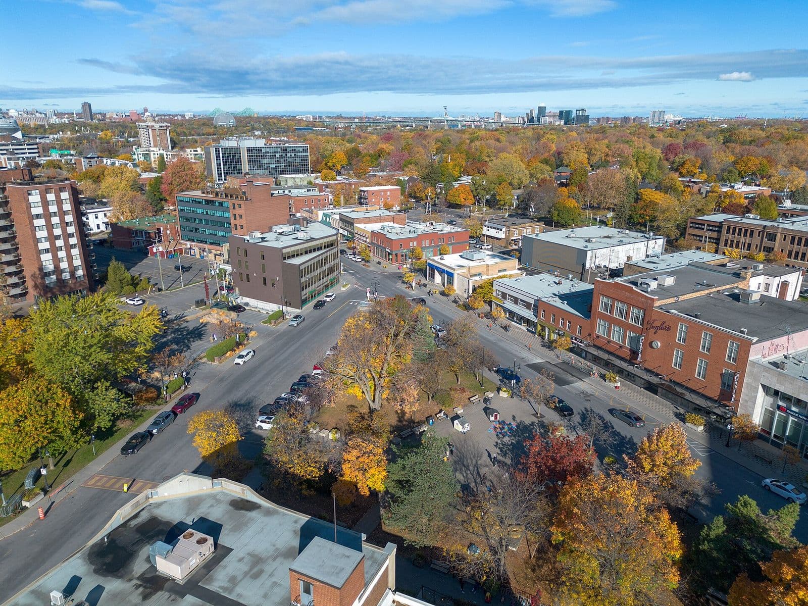 Vue aérienne du village de Saint-Lambert autour de l’avenue Victoria à l’automne, avec ses commerces de brique, un petit square aux arbres orangés et le pont Jacques-Cartier à l’horizon