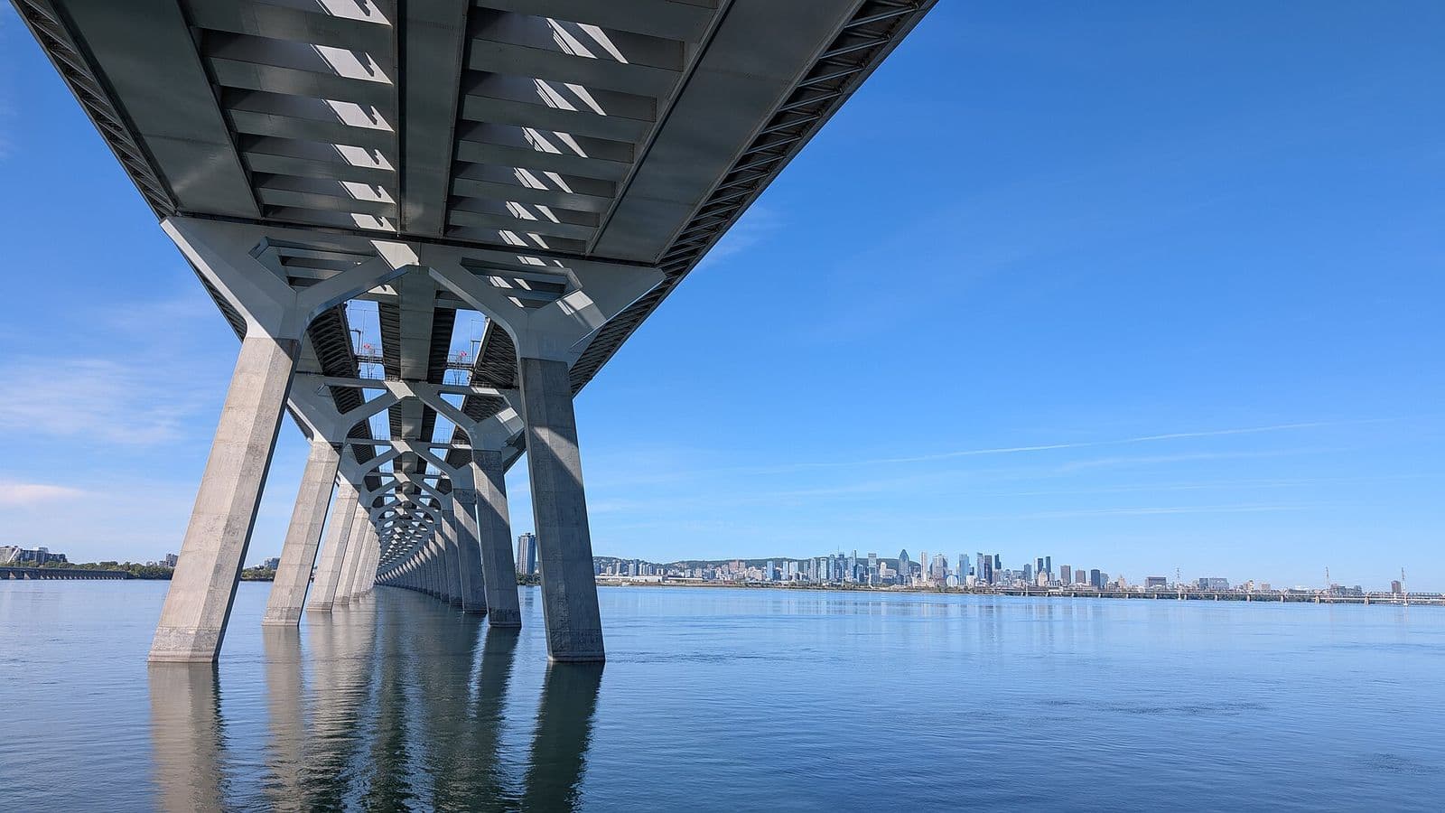 The concrete piers of the Samuel-De Champlain Bridge march across the calm St. Lawrence from Brossard toward the downtown Montréal skyline under a clear blue sky