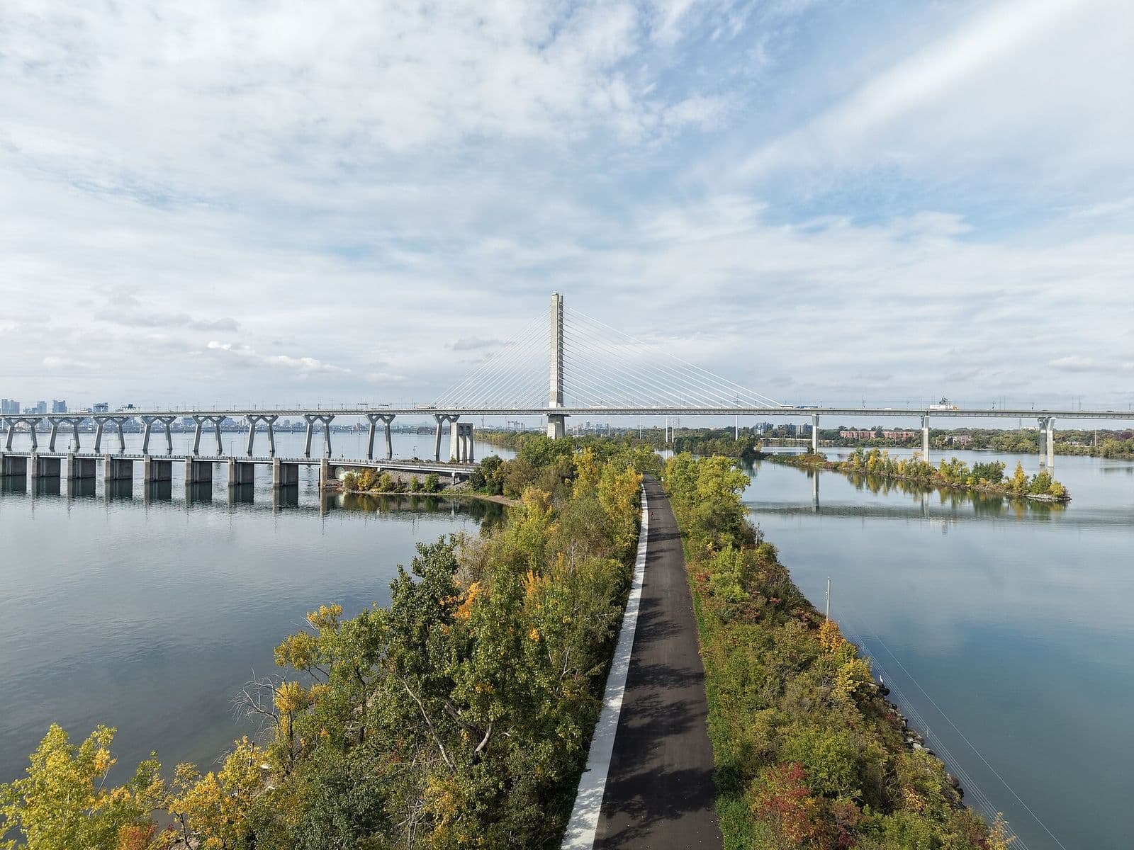 Une piste cyclable asphaltée longe une digue étroite et boisée entre deux plans d’eau calmes, vers le pylône du pont Samuel-De Champlain