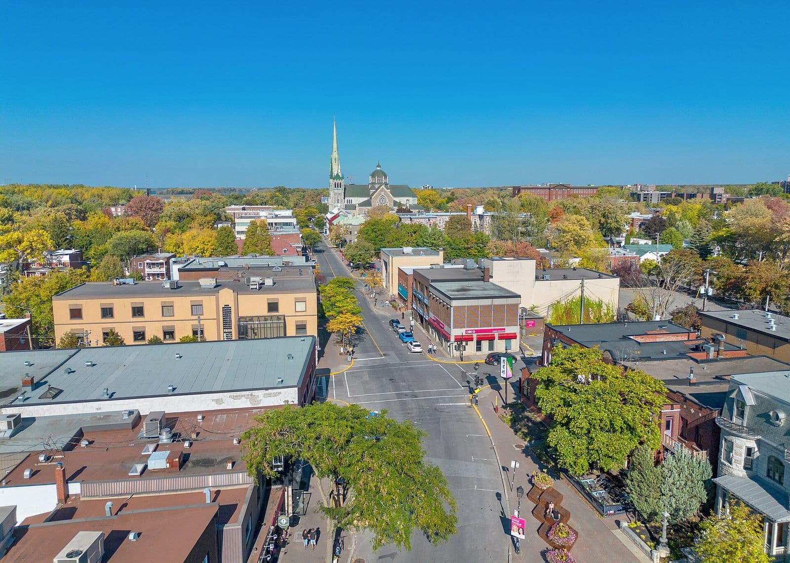 Vue aérienne d’une rue commerçante bordée d’arbres et de bâtiments de brique du Vieux-Longueuil menant à la haute flèche de la cocathédrale Saint-Antoine-de-Padoue, aux premières couleurs de l’automne
