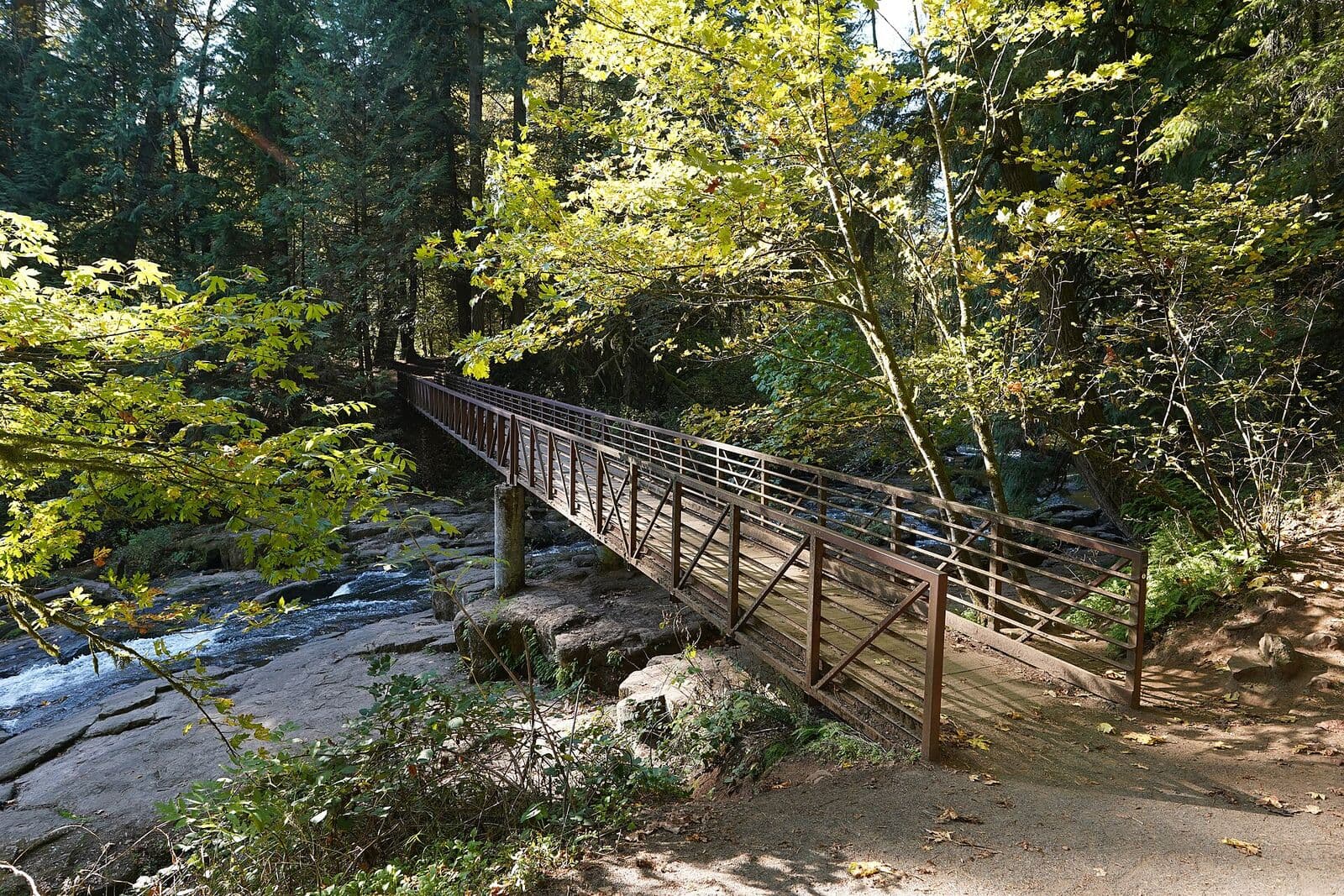 A long steel footbridge crossing Lacamas Creek between rock ledges and sunlit autumn maples on the trail in Camas