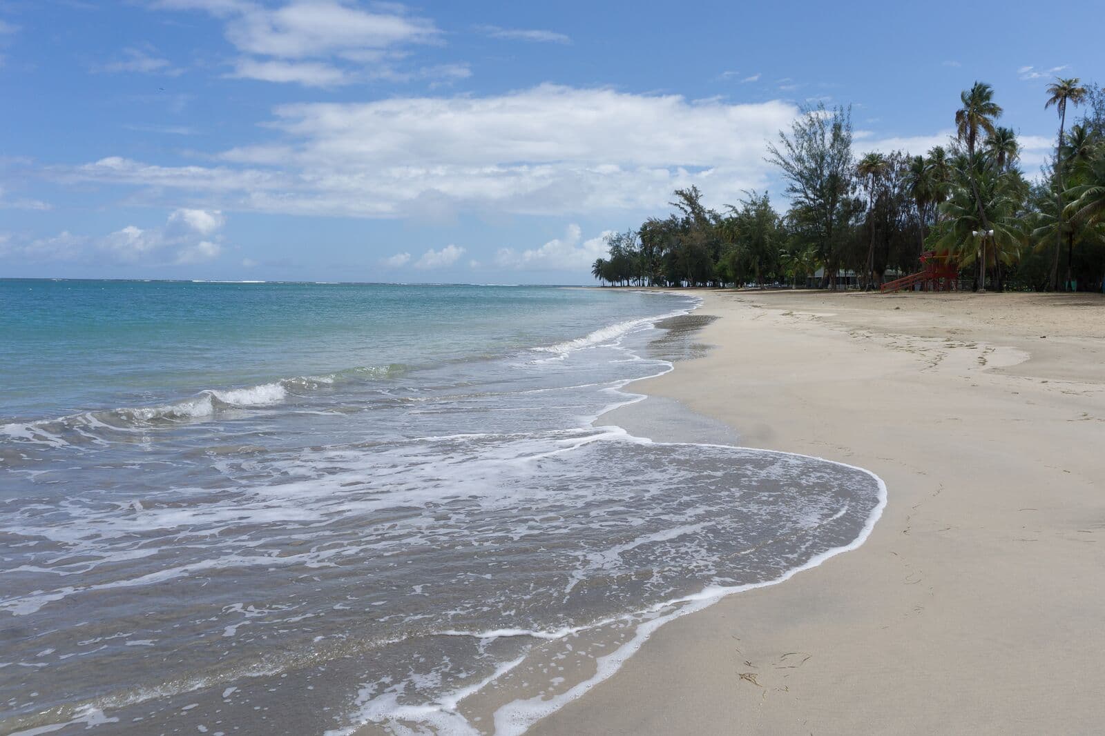 Gentle waves on a wide, empty sand beach lined with palms at Luquillo, a red lifeguard stand in the distance