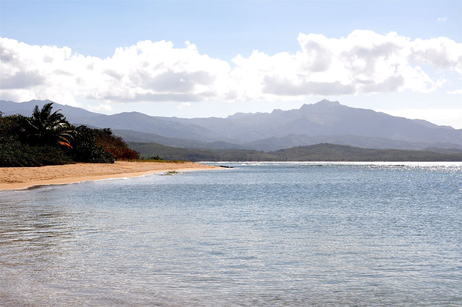 A calm bay and a curving sand beach at Juan Martín, Luquillo, with the El Yunque mountains under white clouds behind