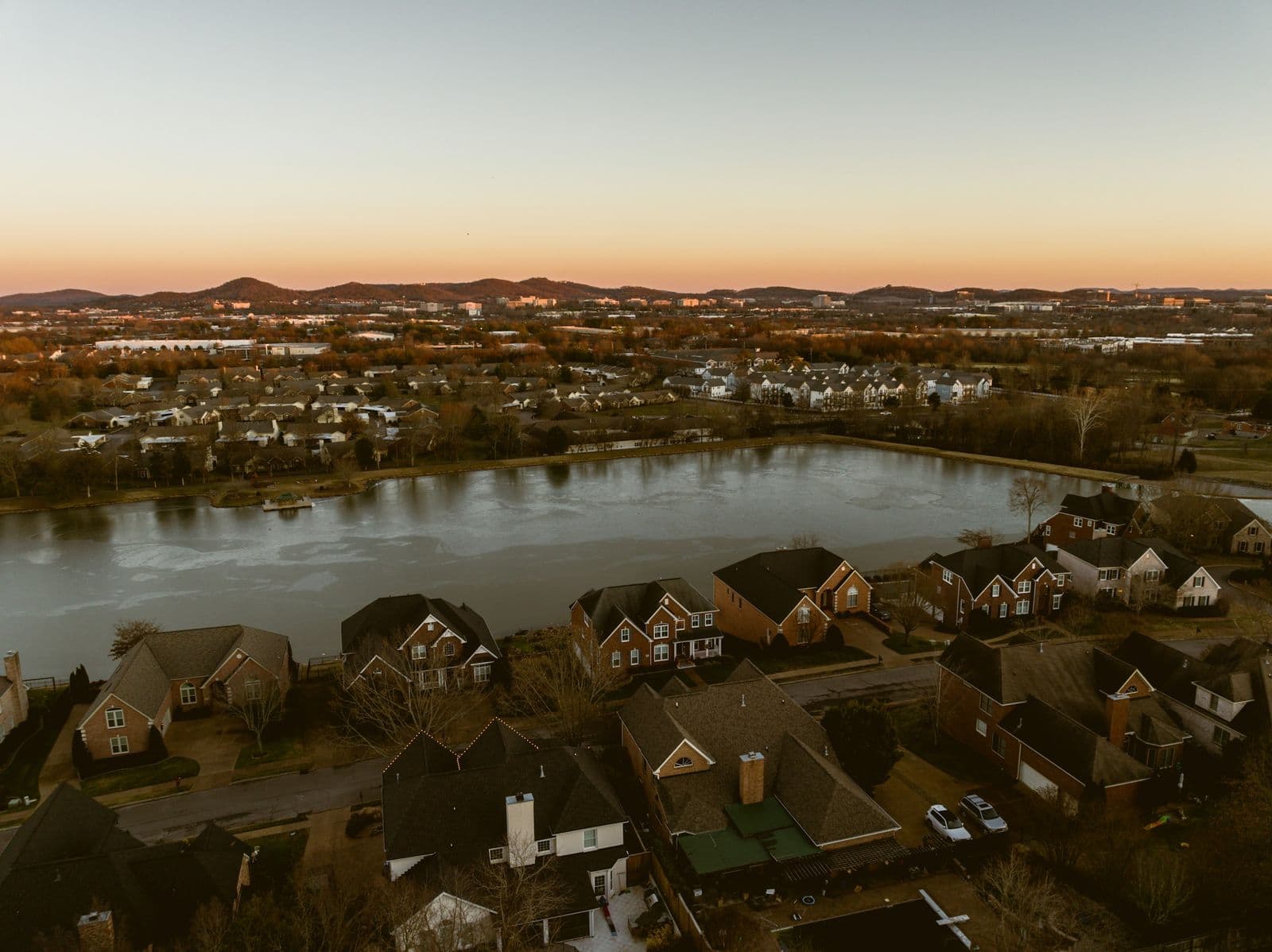 Aerial view at dusk of brick two-story houses around a pond in a Franklin neighborhood, with more neighborhoods and hills beyond