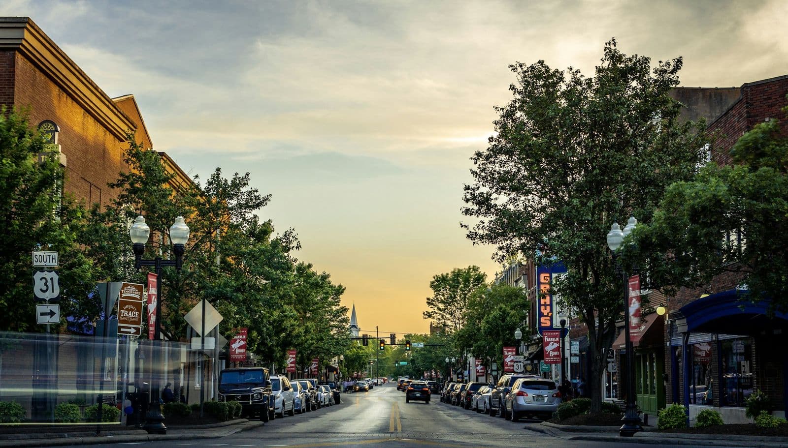 Main Street in downtown Franklin at sunset, with brick storefronts, street trees, lamp posts, Franklin Rodeo banners and a church steeple at the end of the street