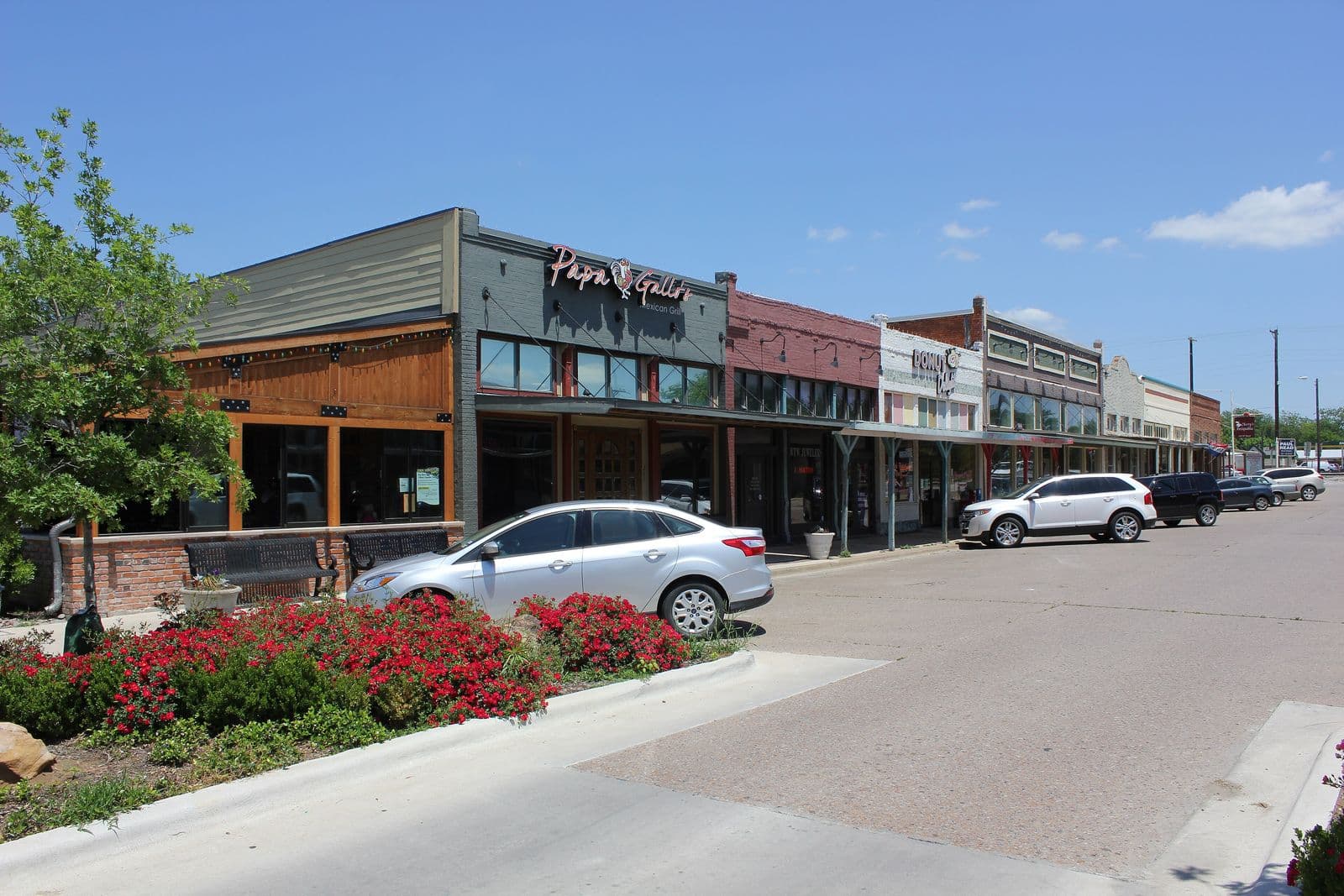 A row of old brick storefronts with covered sidewalks on Celina’s downtown square, red roses in the foreground and a blue sky