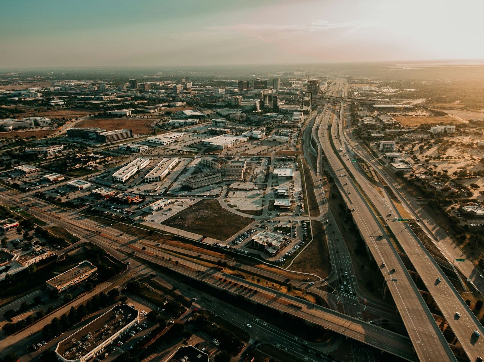 Aerial view of Frisco at golden hour: office towers, new buildings under construction and a wide tollway with flyover ramps running north toward the horizon