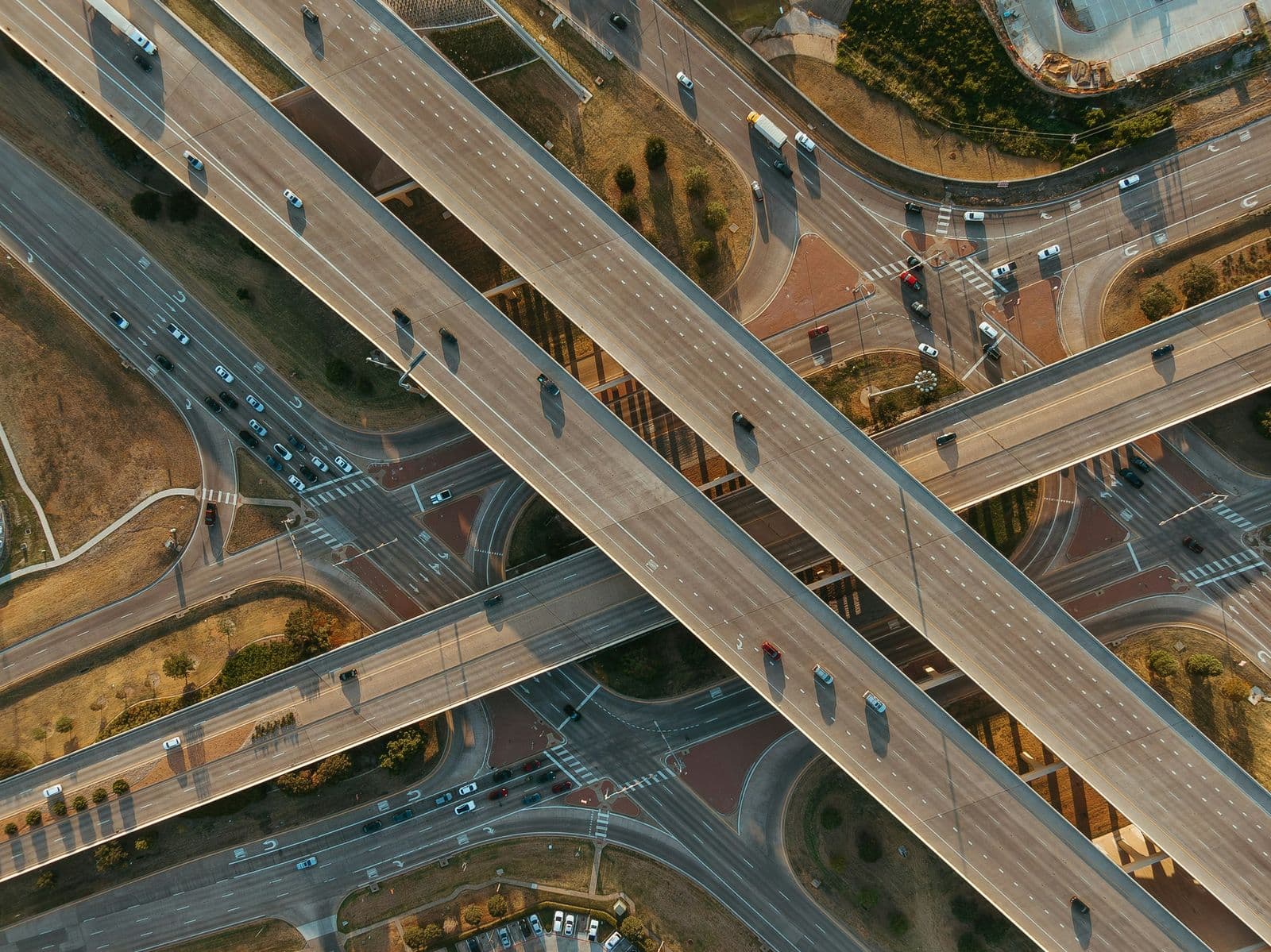 Top-down view of a stacked highway interchange in Frisco in late-afternoon light, elevated lanes crossing over busy frontage roads