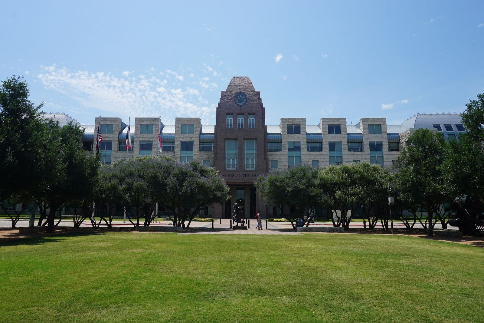 Frisco’s stone municipal center with its clock tower, seen across a green lawn and a row of flowering trees on a clear summer day