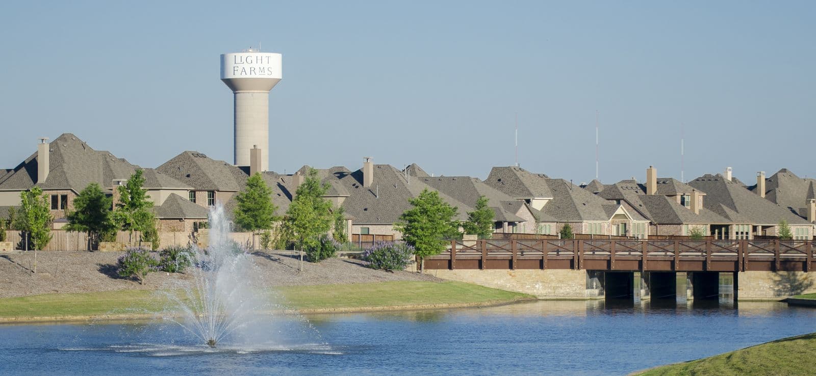 A fountain in a blue lake at Light Farms in Celina, with a footbridge, a water tower and rows of new brick homes behind it