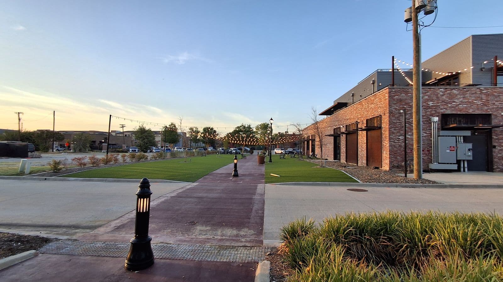 A brick-paved walkway with strings of lights overhead between green lawns and a new red-brick building in Prosper at dusk