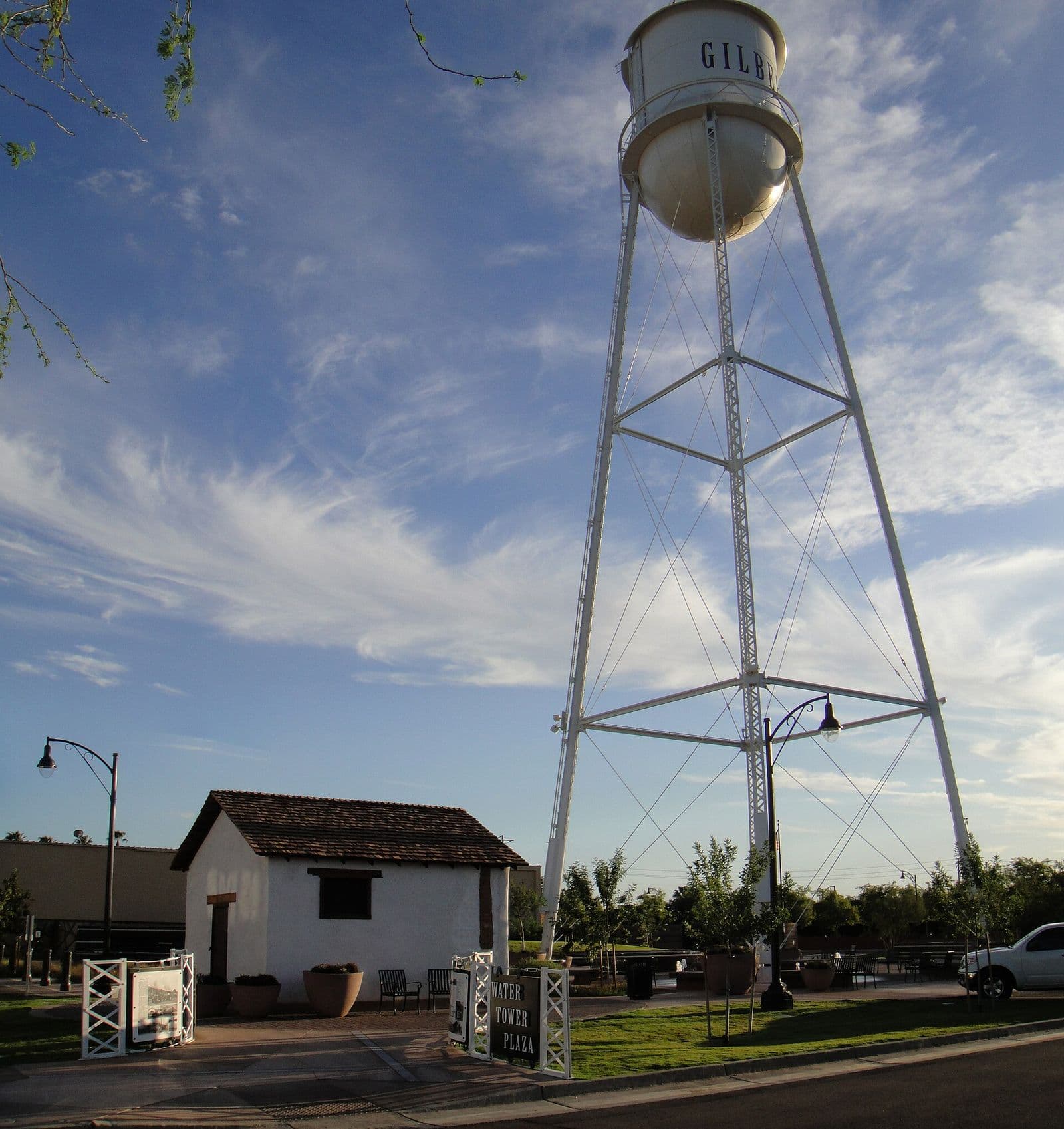 The white Gilbert water tower above Water Tower Plaza in downtown Gilbert, with a small historic white outbuilding and the plaza sign below