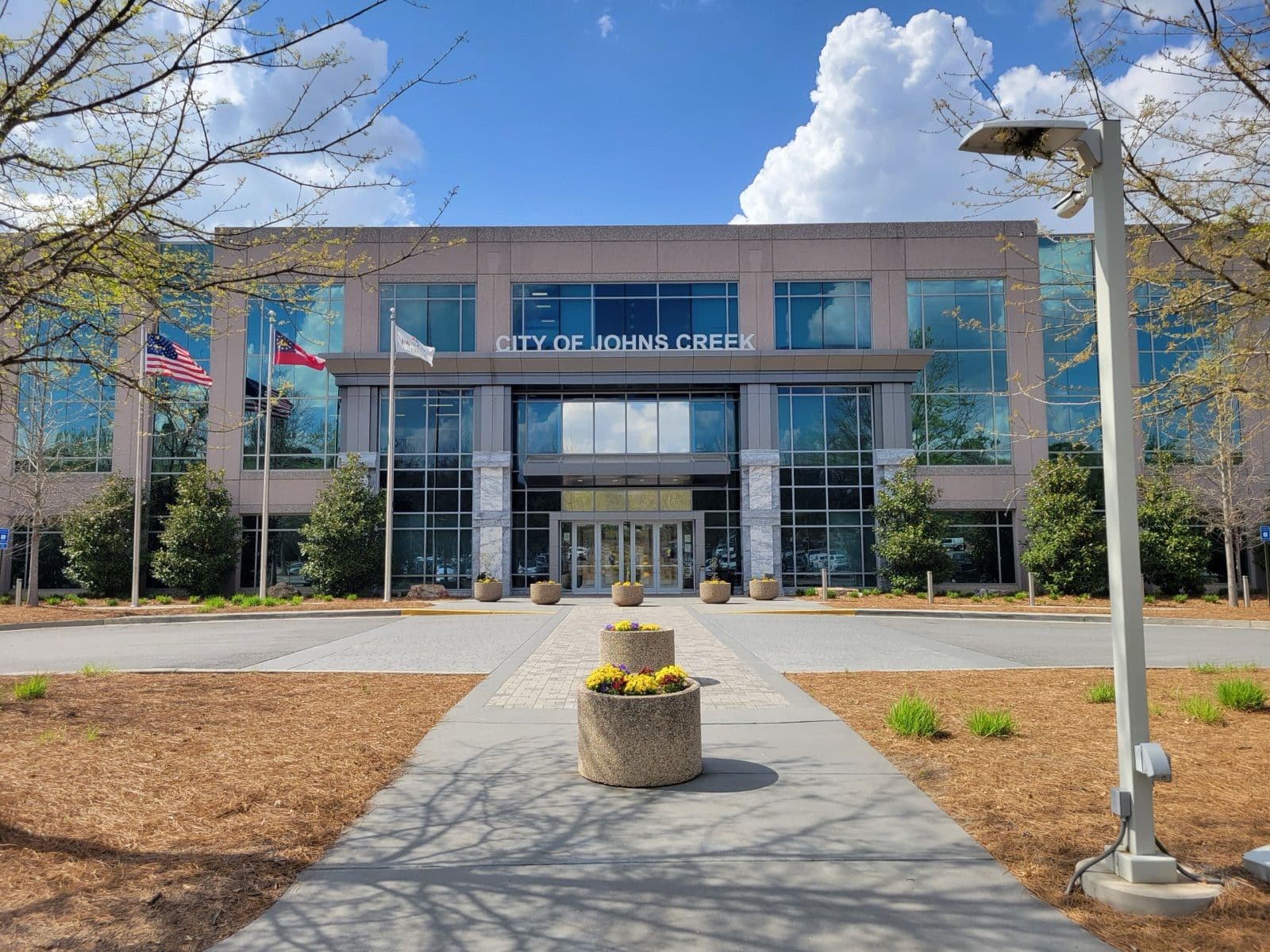 Johns Creek City Hall, a glass-fronted building with flags and spring flower planters, in the Town Center area