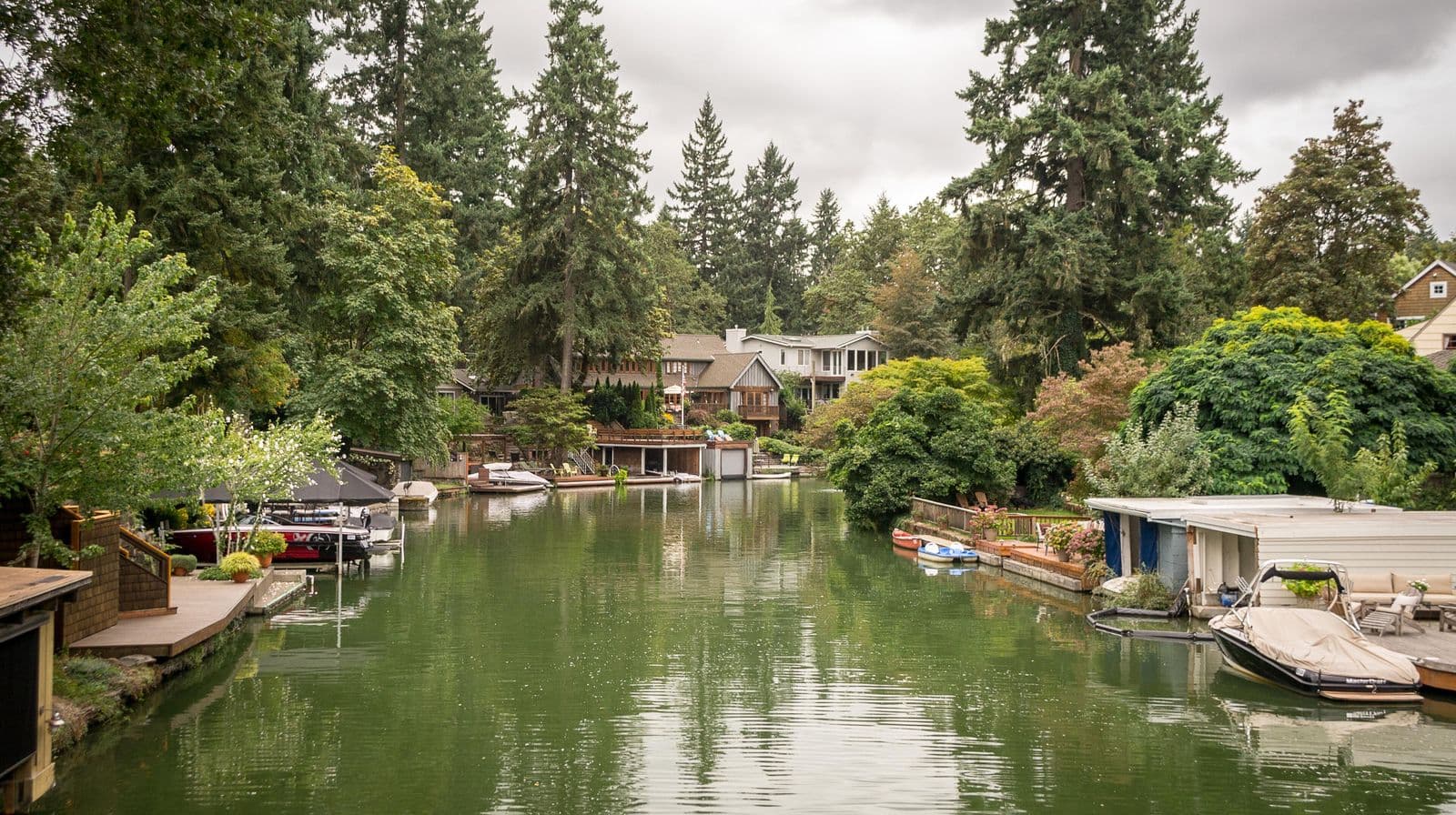 A calm green channel of Oswego Lake lined with boathouses, docks, moored boats and homes under tall Douglas firs
