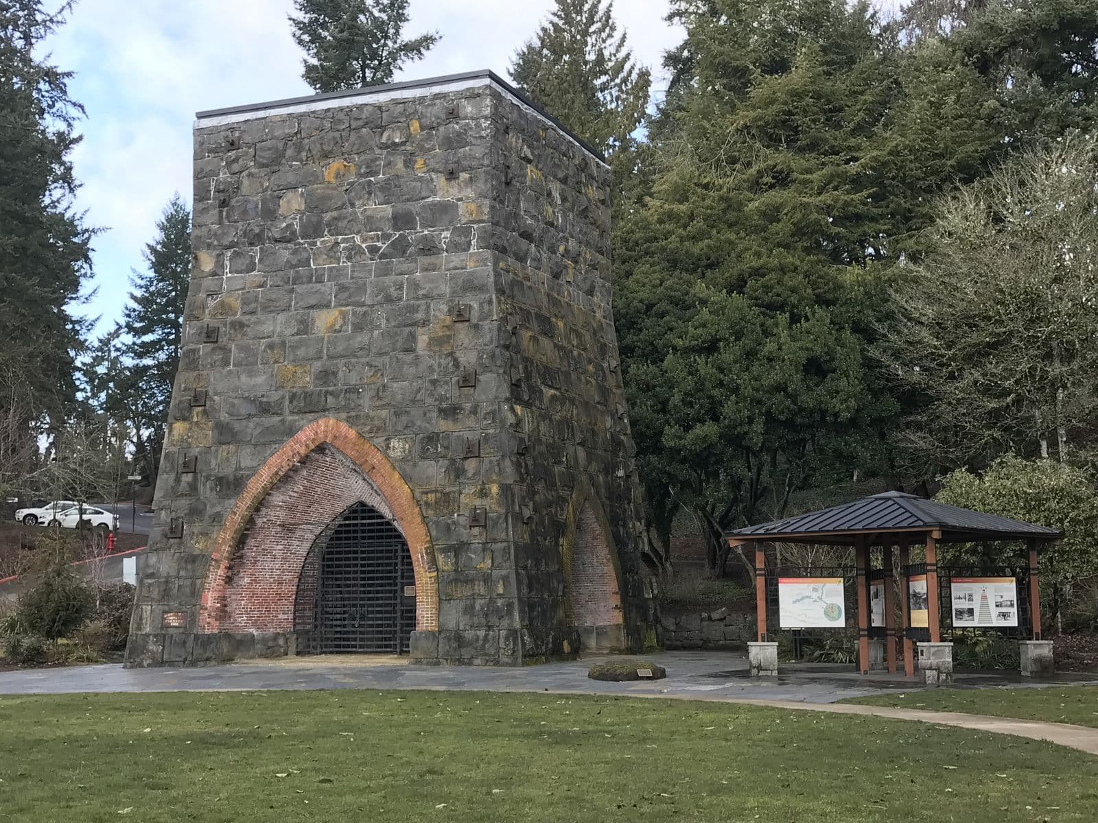 The tapering stone Oregon Iron Company furnace with its red-brick arch, beside a lawn and a small kiosk of history panels in George Rogers Park