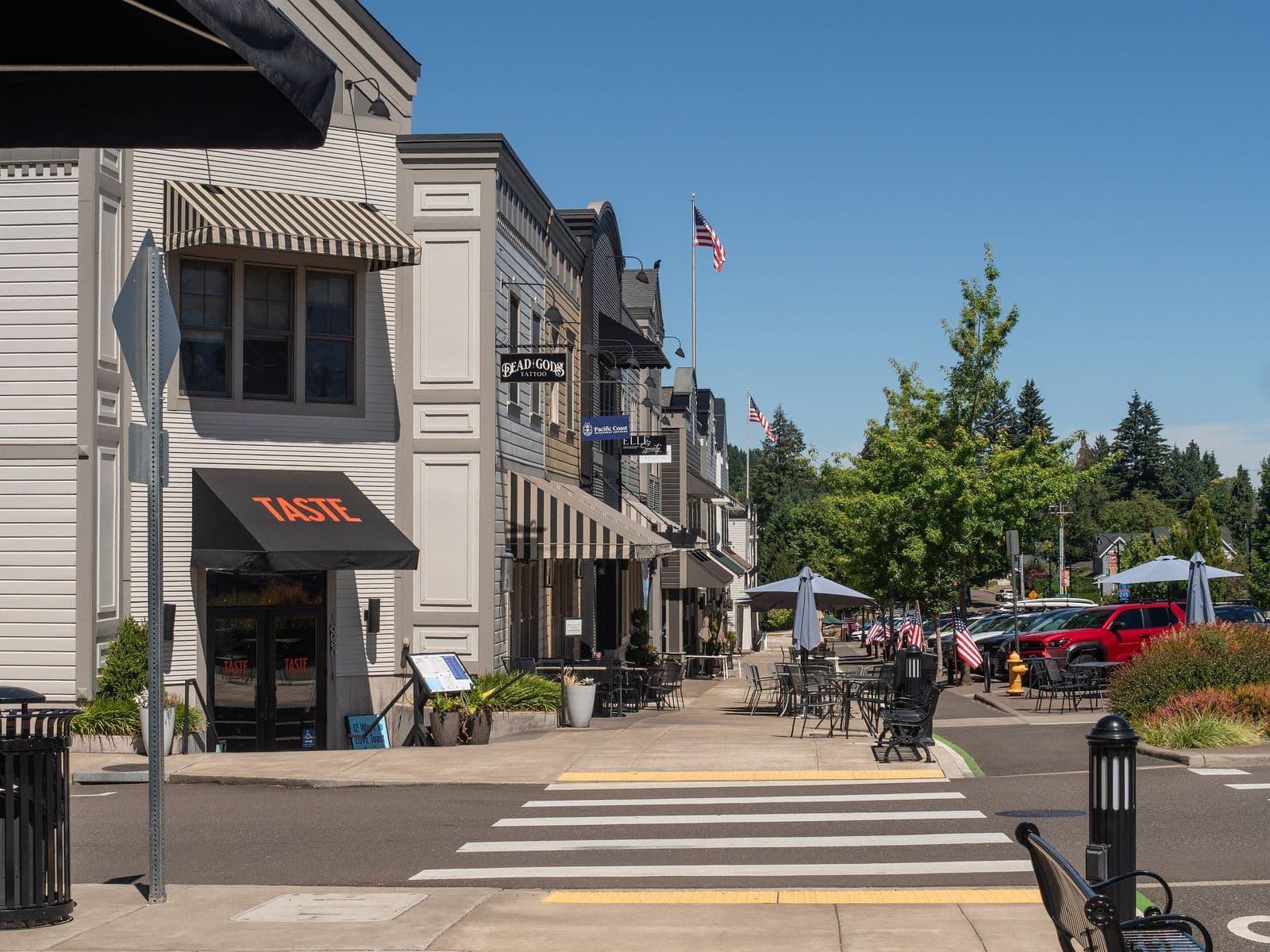 A sunny main street in West Linn’s historic Willamette district, with gray clapboard storefronts, striped awnings, sidewalk tables, flags and a crosswalk