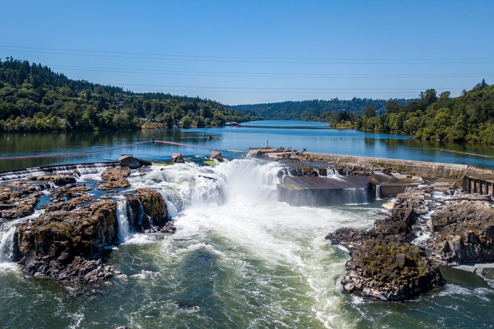 Aerial view of Willamette Falls pouring over dark basalt ledges, with the wide Willamette River and wooded hills stretching upstream under a clear blue sky