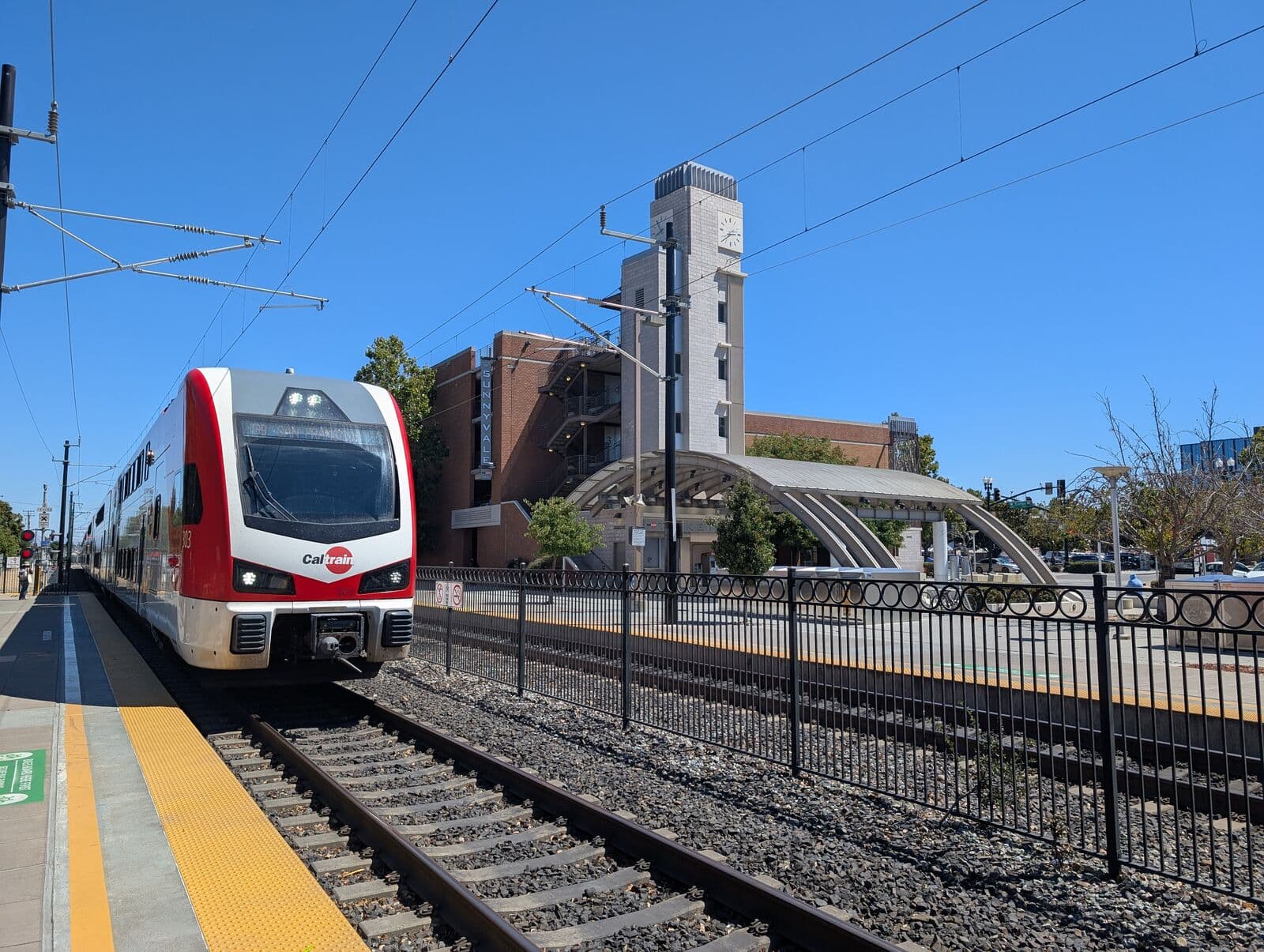 A red and white electric Caltrain train arriving at Sunnyvale station, with the station clock tower and arched shelter under a clear blue sky