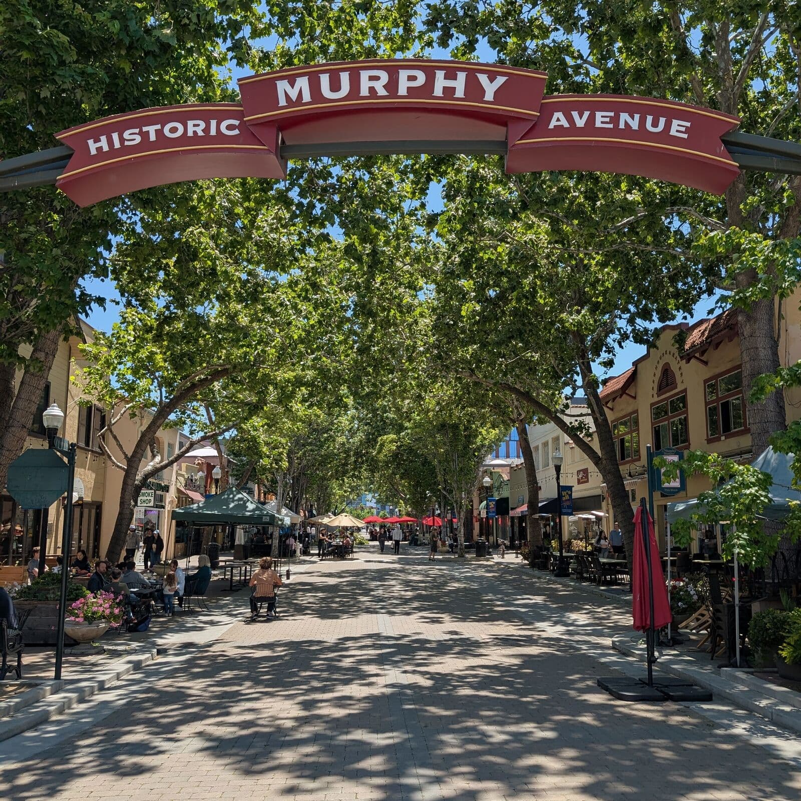 The Historic Murphy Avenue arch over a tree-shaded pedestrian street of low storefronts and café tables in downtown Sunnyvale on a sunny spring day
