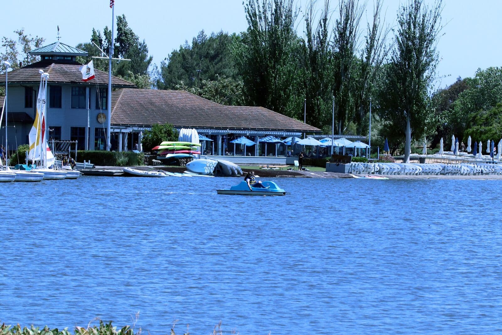 The Shoreline Lake boathouse in Mountain View with sailboats, kayaks and blue umbrellas on the shore and a pedal boat out on the water