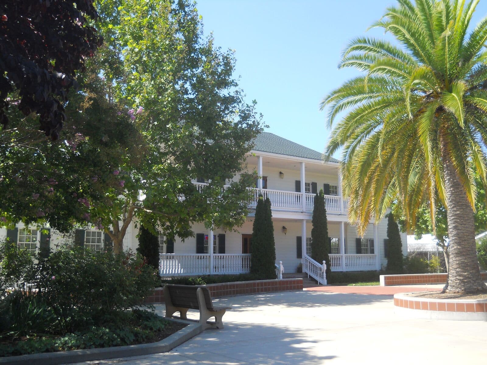 The white two-story Sunnyvale Heritage Park Museum, a replica of the Murphy family home with wide porches, beside a palm tree and a courtyard bench