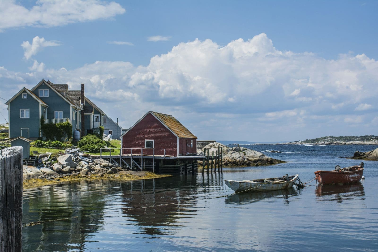 A red fish shed on stilts and a blue shingled house beside a calm cove at Peggy's Cove, with two small boats moored in the water under summer clouds