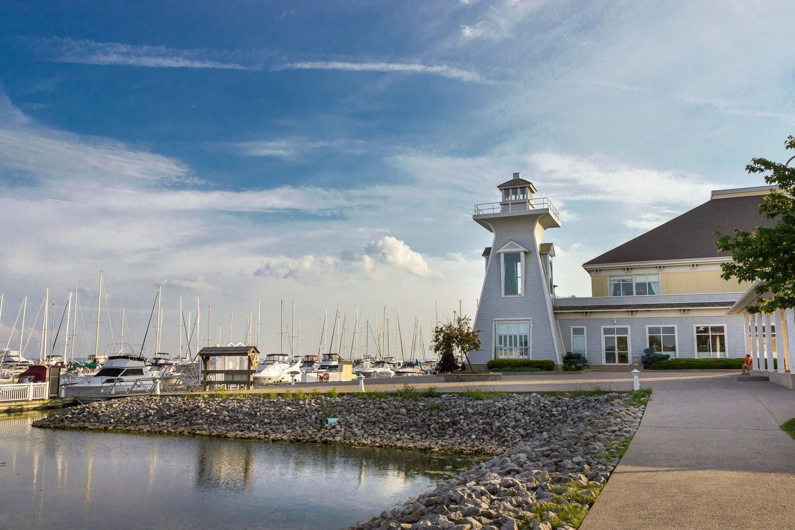 A grey lighthouse-style building beside the marina at Bronte Heritage Waterfront Park in Oakville, with sailboat masts along the docks and a rock-lined shore