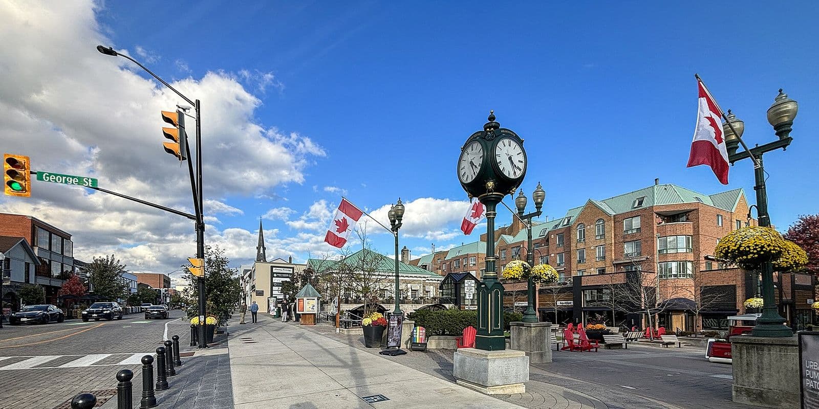Lakeshore Road East at George Street in downtown Oakville, with a green post clock, Canadian flags, hanging flower baskets and red-brick shops under a blue sky