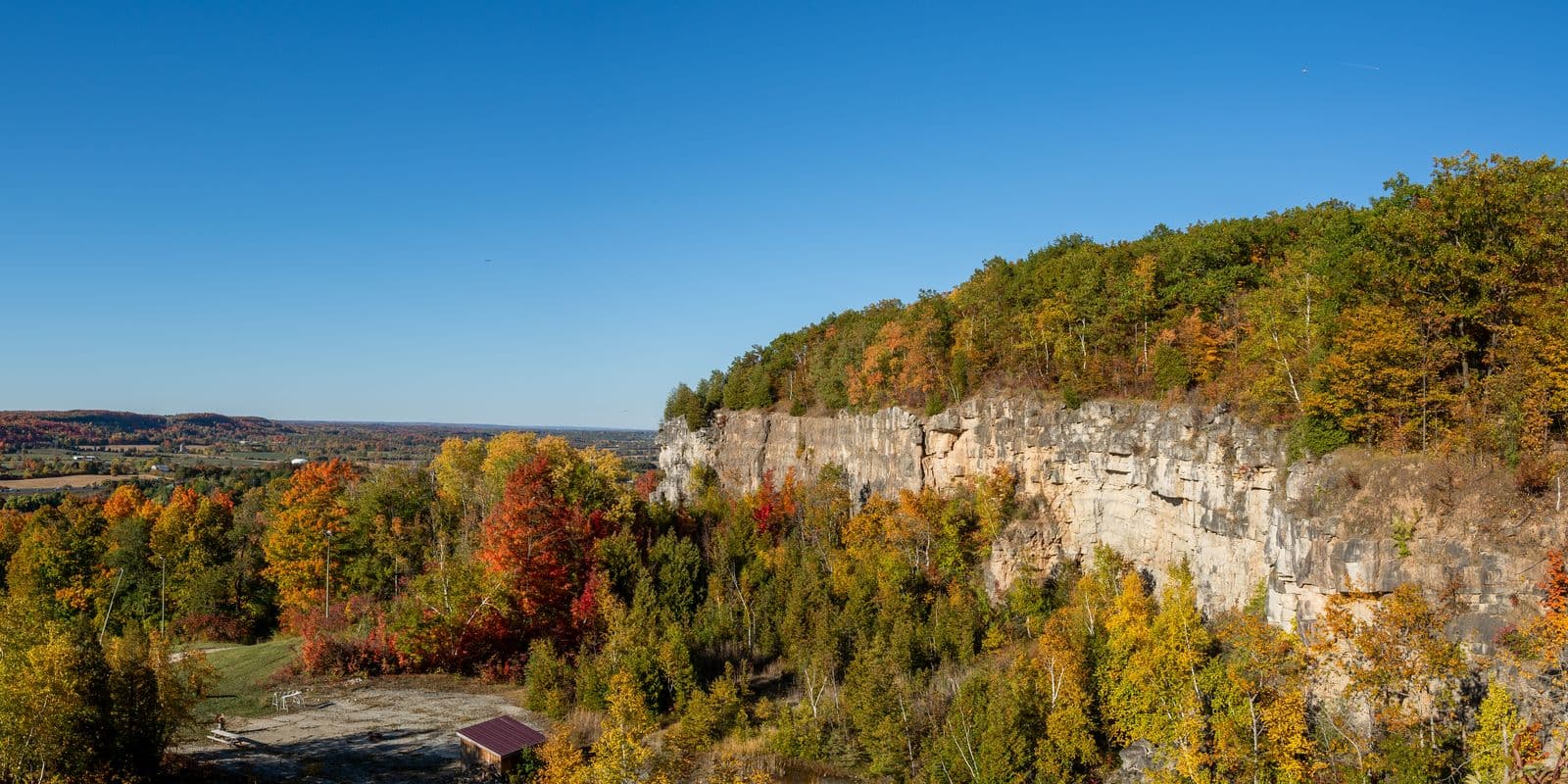 Limestone cliffs of the Niagara Escarpment above a forest in full autumn colour at Kelso Conservation Area, with farmland stretching to the horizon