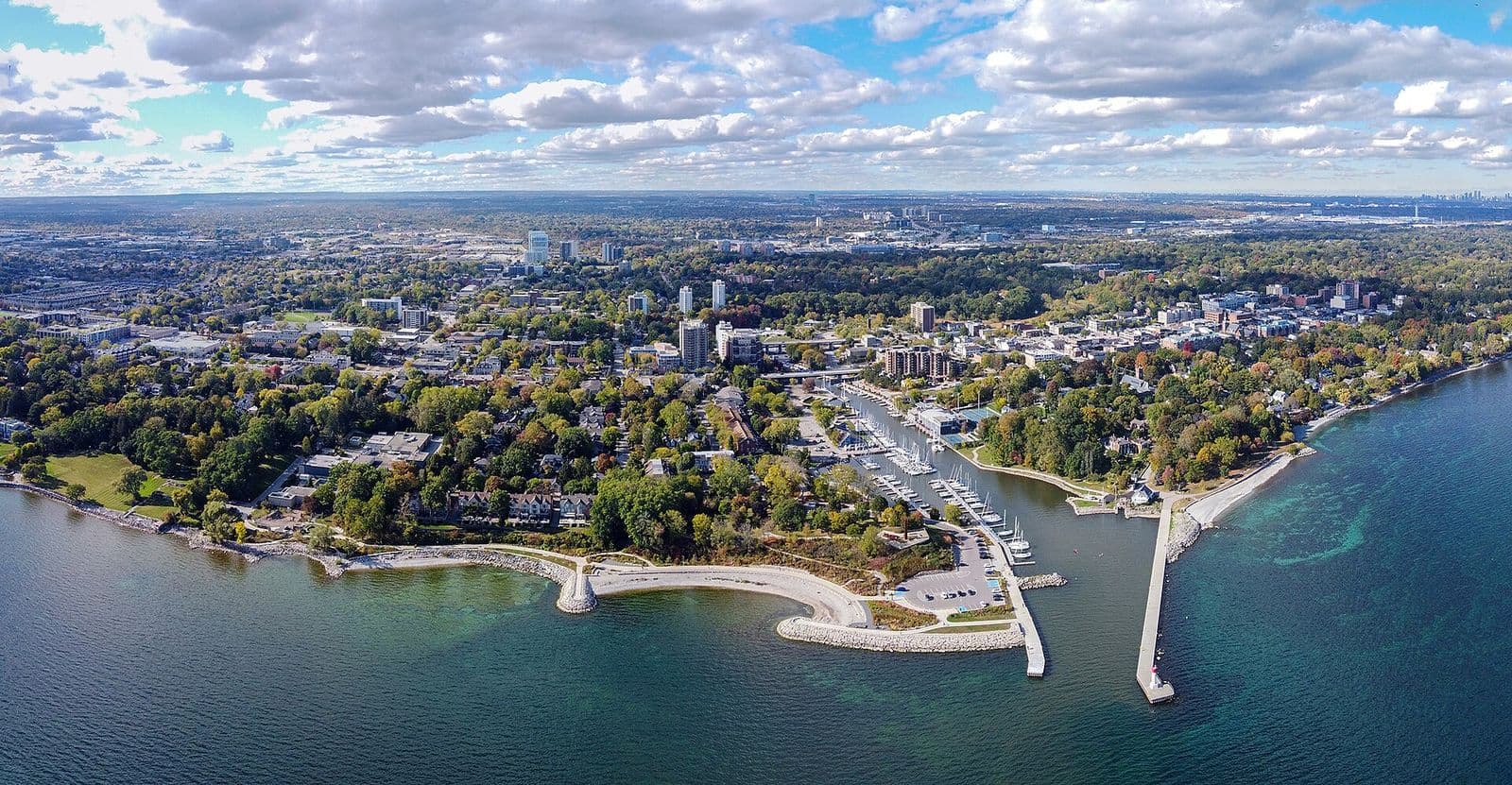 Aerial view of Oakville Harbour at the mouth of Sixteen Mile Creek, with sailboats along the creek, a curving stone breakwater and pier on Lake Ontario, and downtown Oakville among autumn trees