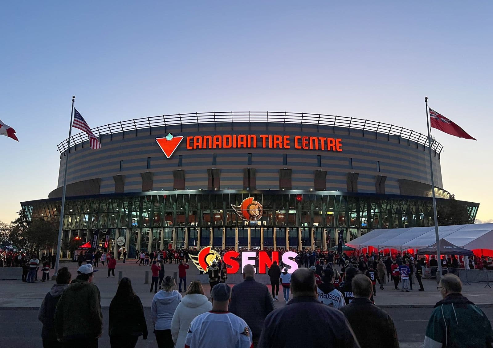 Des partisans marchent vers le Centre Canadian Tire illuminé au crépuscule avant un match des Sénateurs d’Ottawa, des drapeaux flottant de chaque côté