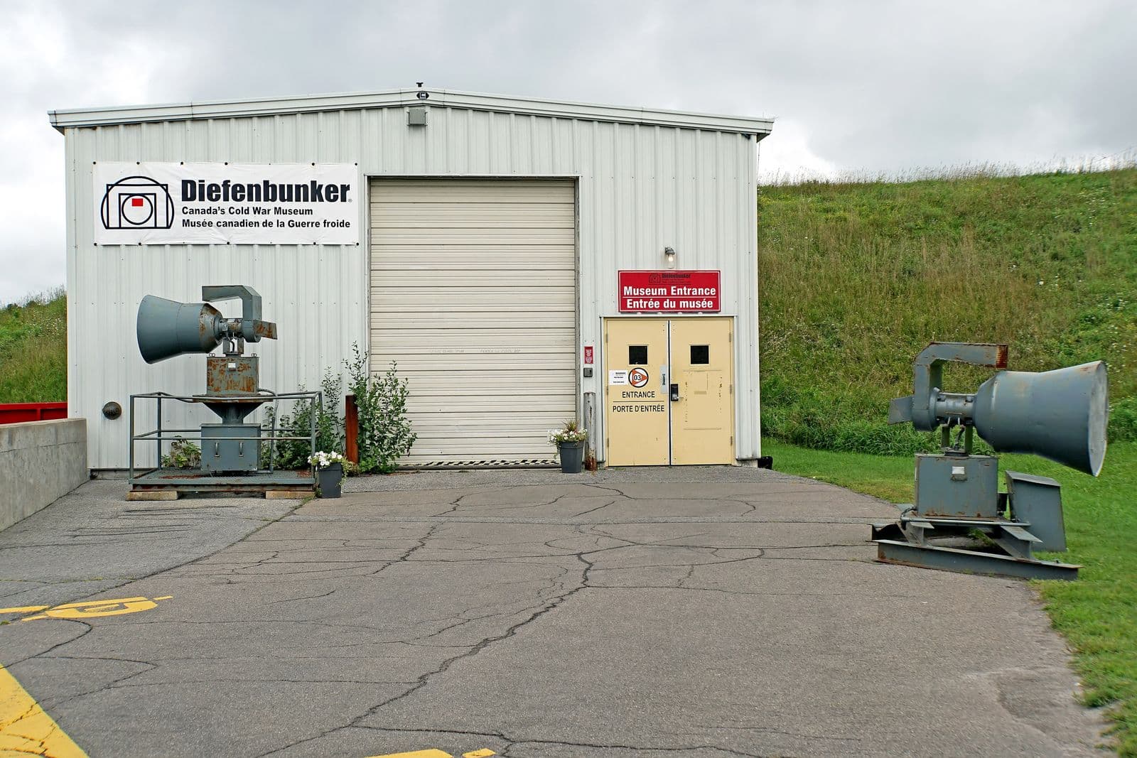 Le bâtiment d’entrée du Diefenbunker, à Carp, avec son enseigne bilingue de musée et deux sirènes d’alerte aérienne de la guerre froide le long de l’allée